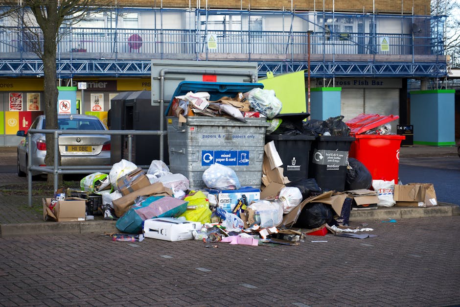 A large pile of black plastic rubbish bags, some tied tightly and others loosely sealed, stacked against a modern building wall with a tiled surface. The bags contain various waste items, including vi